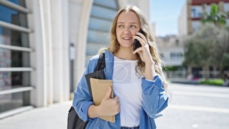 Téléchargez les photos : Jeune étudiante blonde tenant des livres parlant sur smartphone à l'université - en image libre de droit