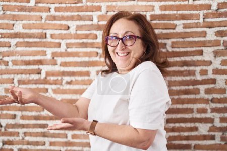 Téléchargez les photos : Femme âgée avec des lunettes debout sur le mur de briques invitant à entrer souriant naturel avec la main ouverte - en image libre de droit