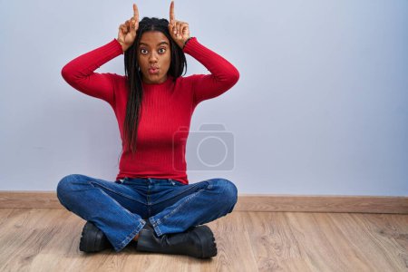 Foto de Joven afroamericano con trenzas sentado en el suelo en casa haciendo un gesto divertido con el dedo sobre la cabeza como cuernos de toro - Imagen libre de derechos