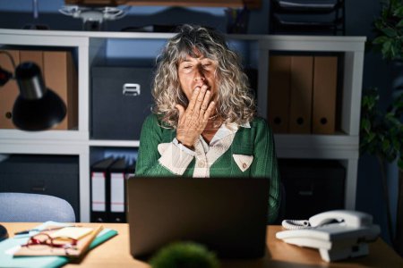 Téléchargez les photos : Femme d'âge moyen travaillant la nuit à l'aide d'un ordinateur portable ennuyé bâillement fatigué couvrant la bouche avec la main. agitation et somnolence. - en image libre de droit
