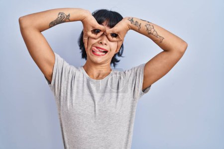 Photo for Young hispanic woman wearing casual t shirt over blue background doing ok gesture like binoculars sticking tongue out, eyes looking through fingers. crazy expression. - Royalty Free Image