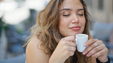 Photo for Young beautiful hispanic woman sitting on table smelling cup of coffee smiling at coffee shop terrace - Royalty Free Image