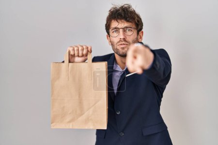 Téléchargez les photos : Homme d'affaires hispanique tenant sac de livraison pointant du doigt la caméra et à vous, geste confiant regardant sérieux - en image libre de droit