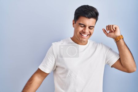 Foto de Hombre hispano de pie sobre fondo azul bailando feliz y alegre, sonriendo moviéndose casual y confiado escuchando música - Imagen libre de derechos