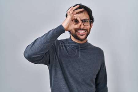 Photo for Handsome latin man standing over isolated background doing ok gesture with hand smiling, eye looking through fingers with happy face. - Royalty Free Image