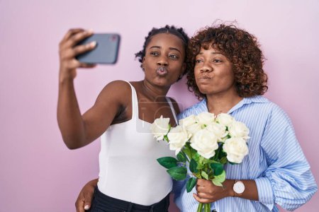 Foto de Dos mujeres africanas tomando una foto selfie con flores hinchando mejillas con cara divertida. boca inflada con aire, captación de aire. - Imagen libre de derechos