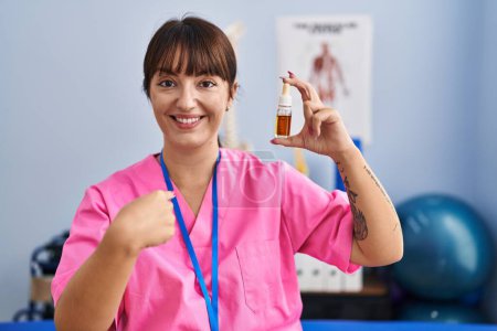 Téléchargez les photos : Jeune femme brune tenant l'huile cbd à la clinique de physiothérapie pointant du doigt un sourire heureux et fier - en image libre de droit