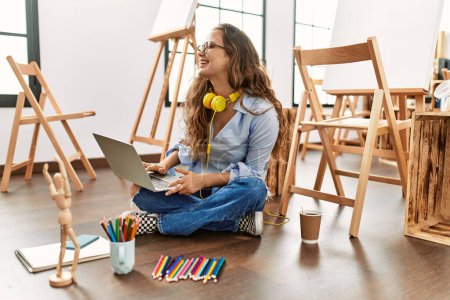 Téléchargez les photos : Jeune belle artiste hispanique femme en utilisant ordinateur portable assis sur le sol au studio d'art - en image libre de droit