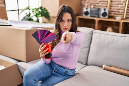 Photo for Young brunette woman moving to a new home choosing wall painting pointing with finger to the camera and to you, confident gesture looking serious - Royalty Free Image