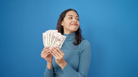 Foto de Joven hermosa mujer hispana sonriendo confiada sosteniendo dinero sobre aislado fondo azul - Imagen libre de derechos