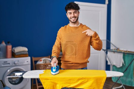 Foto de Hombre hispano con barba planchando ropa en casa señalando con el dedo a uno mismo sonriendo feliz y orgulloso - Imagen libre de derechos