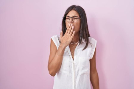 Foto de Brunette young woman standing over pink background wearing glasses bored yawning tired covering mouth with hand. restless and sleepiness. - Imagen libre de derechos