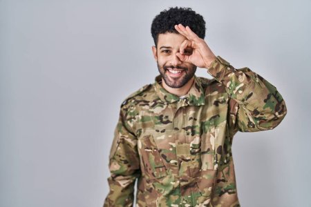Téléchargez les photos : Homme arabe portant un uniforme de camouflage de l'armée faisant un geste correct avec la main souriante, les yeux regardant à travers les doigts avec un visage heureux. - en image libre de droit