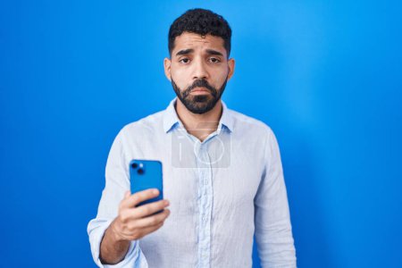 Téléchargez les photos : Hispanic man with beard using smartphone typing message depressed and worry for distress, crying angry and afraid. sad expression. - en image libre de droit