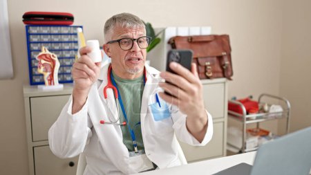 Téléchargez les photos : Moyen âge homme aux cheveux gris médecin ayant une consultation médicale en ligne à la clinique - en image libre de droit