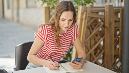 Photo for Young blonde woman sitting on table writing on notebook using smartphone at coffee shop terrace - Royalty Free Image