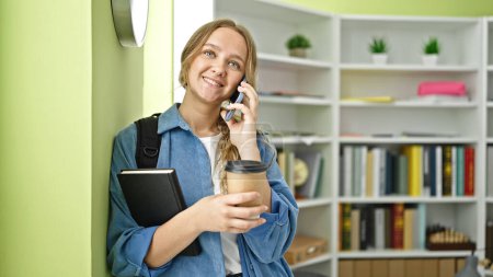 Téléchargez les photos : Jeune étudiante blonde parlant sur smartphone buvant du café à la bibliothèque de l'université - en image libre de droit