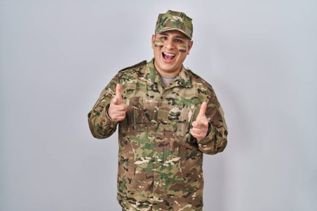Téléchargez les photos : Jeune homme hispanique portant uniforme de camouflage de l'armée pointant les doigts vers la caméra avec un visage heureux et drôle. bonne énergie et vibes. - en image libre de droit