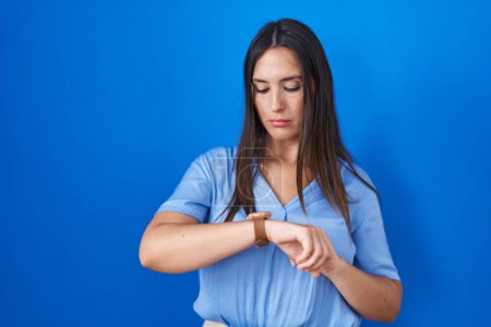 Foto de Mujer morena joven de pie sobre fondo azul comprobar la hora en reloj de pulsera, relajado y seguro de sí mismo - Imagen libre de derechos