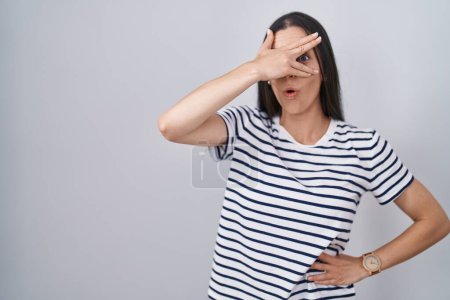 Foto de Joven morena vistiendo camiseta a rayas asomándose en shock cubriendo cara y ojos con la mano, mirando a través de los dedos con expresión avergonzada. - Imagen libre de derechos