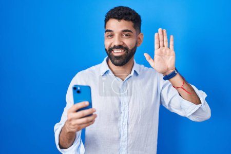 Foto de Hombre hispano con barba usando mensaje de escritura de teléfonos inteligentes renunciando a decir hola feliz y sonriente, gesto de bienvenida amistoso - Imagen libre de derechos