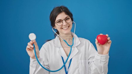 Téléchargez les photos : Jeune belle femme hispanique médecin tenant stéthoscope et coeur sur fond bleu isolé - en image libre de droit