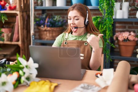 Photo for Redhead woman working at florist shop doing video call pointing thumb up to the side smiling happy with open mouth - Royalty Free Image