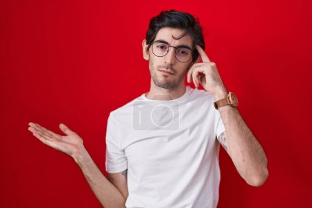 Photo for Young hispanic man standing over red background confused and annoyed with open palm showing copy space and pointing finger to forehead. think about it. - Royalty Free Image