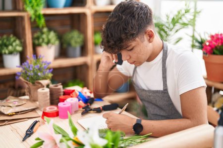 Photo for Young hispanic teenager florist talking on smartphone writing on notebook at flower shop - Royalty Free Image