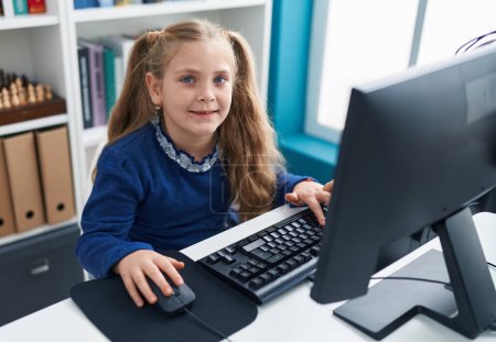 Téléchargez les photos : Adorable étudiante blonde utilisant un ordinateur assis sur la table en classe - en image libre de droit