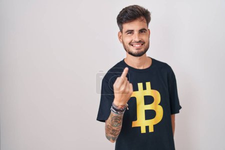 Foto de Joven hombre hispano con tatuajes con camiseta bitcoin haciendo señas ven aquí gesto con la mano invitando a la bienvenida feliz y sonriente - Imagen libre de derechos