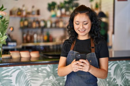 Téléchargez les photos : Young chinese woman waitress smiling confident using smartphone at restaurant - en image libre de droit