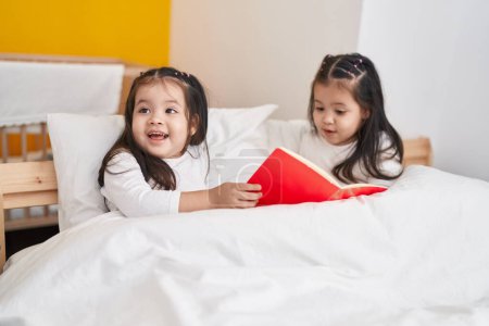 Photo for Adorable twin girls reading book sitting on bed at bedroom - Royalty Free Image