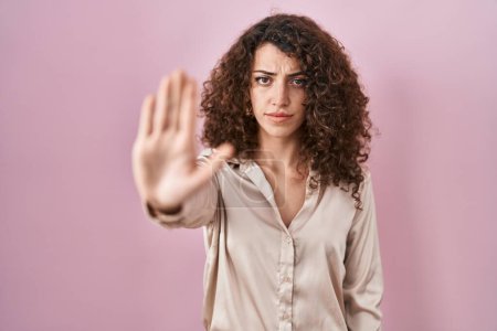 Foto de Mujer hispana con el pelo rizado de pie sobre fondo rosa haciendo dejar de cantar con la palma de la mano. expresión de advertencia con gesto negativo y grave en la cara. - Imagen libre de derechos