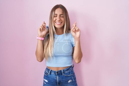 Téléchargez les photos : Young blonde woman standing over pink background gesturing finger crossed smiling with hope and eyes closed. luck and superstitious concept. - en image libre de droit
