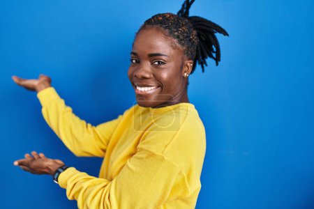 Foto de Beautiful black woman standing over blue background inviting to enter smiling natural with open hand - Imagen libre de derechos