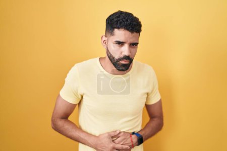 Téléchargez les photos : Hispanic man with beard standing over yellow background with hand on stomach because indigestion, painful illness feeling unwell. ache concept. - en image libre de droit