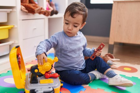 Téléchargez les photos : Adorable garçon hispanique jouant avec des outils jouet assis sur le sol à la maternelle - en image libre de droit