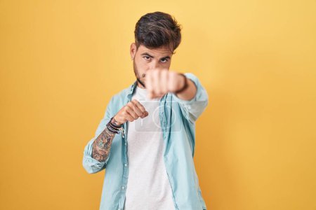 Téléchargez les photos : Jeune homme hispanique avec des tatouages debout sur fond jaune coups de poing poing pour se battre, attaque agressive et en colère, menace et violence - en image libre de droit