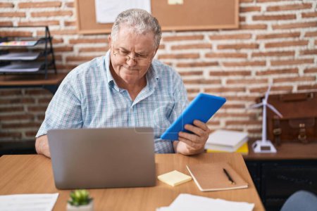 Téléchargez les photos : Homme d'affaires aux cheveux gris d'âge moyen utilisant un pavé tactile et un ordinateur portable au bureau - en image libre de droit