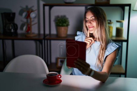 Téléchargez les photos : Jeune femme hispanique utilisant pavé tactile assis sur la table la nuit en pensant concentré sur le doute avec le doigt sur le menton et la recherche jusqu'à se demander - en image libre de droit