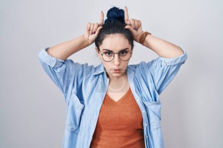 Téléchargez les photos : Jeune fille moderne avec les cheveux bleus debout sur fond blanc faisant geste drôle avec le doigt sur la tête comme cornes de taureau - en image libre de droit