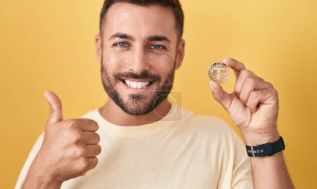 Foto de Hombre hispano guapo sosteniendo la moneda criptomoneda atadura sonriendo feliz y positivo, pulgar hacia arriba haciendo excelente y signo de aprobación - Imagen libre de derechos