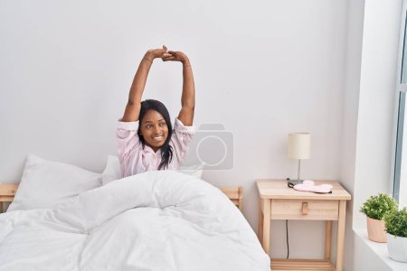 Photo for Young african american woman stretching arms sitting on bed at bedroom - Royalty Free Image