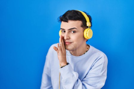 Photo for Non binary person listening to music using headphones hand on mouth telling secret rumor, whispering malicious talk conversation - Royalty Free Image
