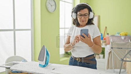 Foto de Mujer hispana de mediana edad escuchando música usando smartphone en la lavandería - Imagen libre de derechos