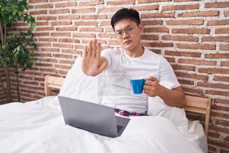 Photo for Young asian man drinking coffee sitting on the bed with open hand doing stop sign with serious and confident expression, defense gesture - Royalty Free Image