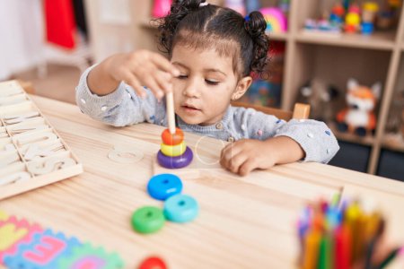 Foto de Adorable chica hispana jugando con aros juguetes sentados en la mesa en el jardín de infantes - Imagen libre de derechos