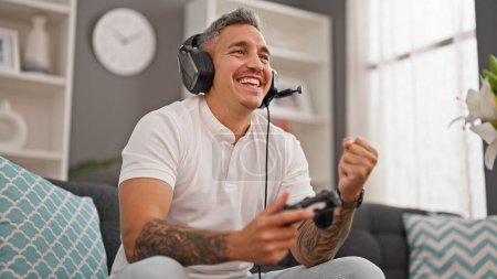 Téléchargez les photos : Jeune homme hispanique jouant à un jeu vidéo assis sur le canapé célébrant à la maison - en image libre de droit