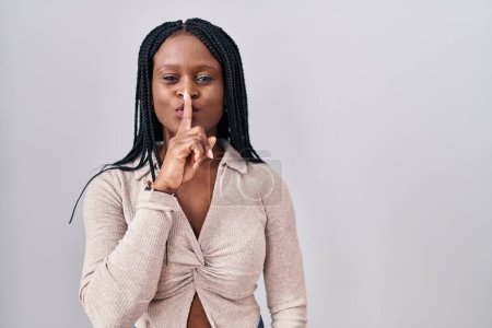 Téléchargez les photos : Femme africaine avec des tresses debout sur fond blanc demandant à être calme avec le doigt sur les lèvres. silence et concept secret. - en image libre de droit